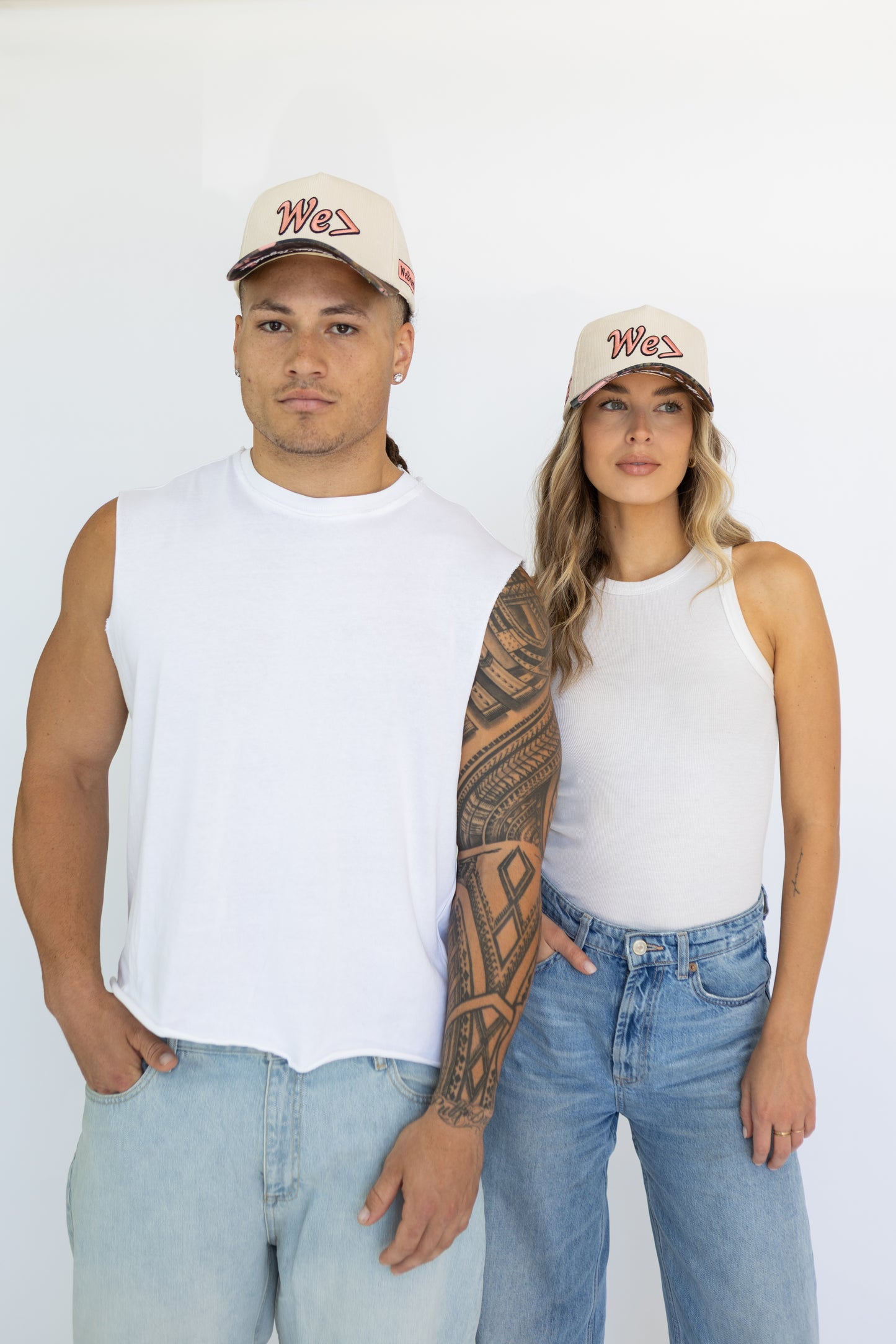 Two people wearing matching white sleeveless shirts and light blue jeans with caps on a white background