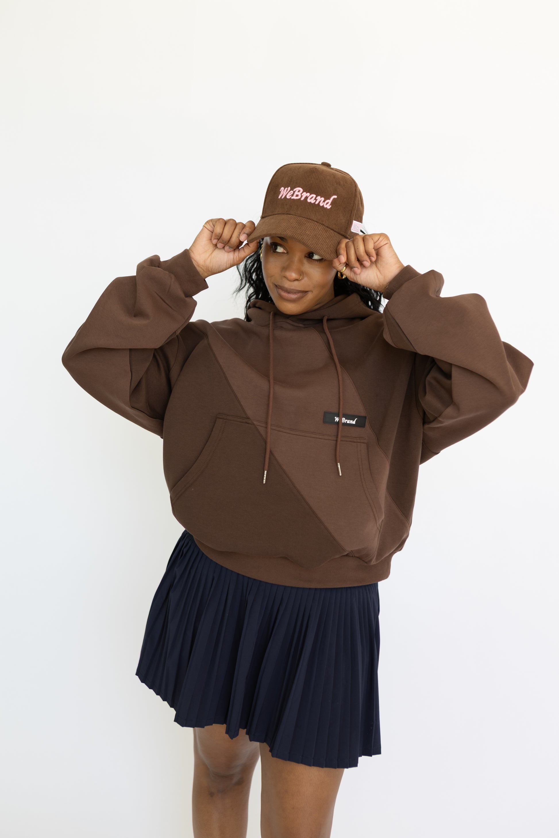 Person wearing a brown hoodie and cap with a navy skirt on a white background