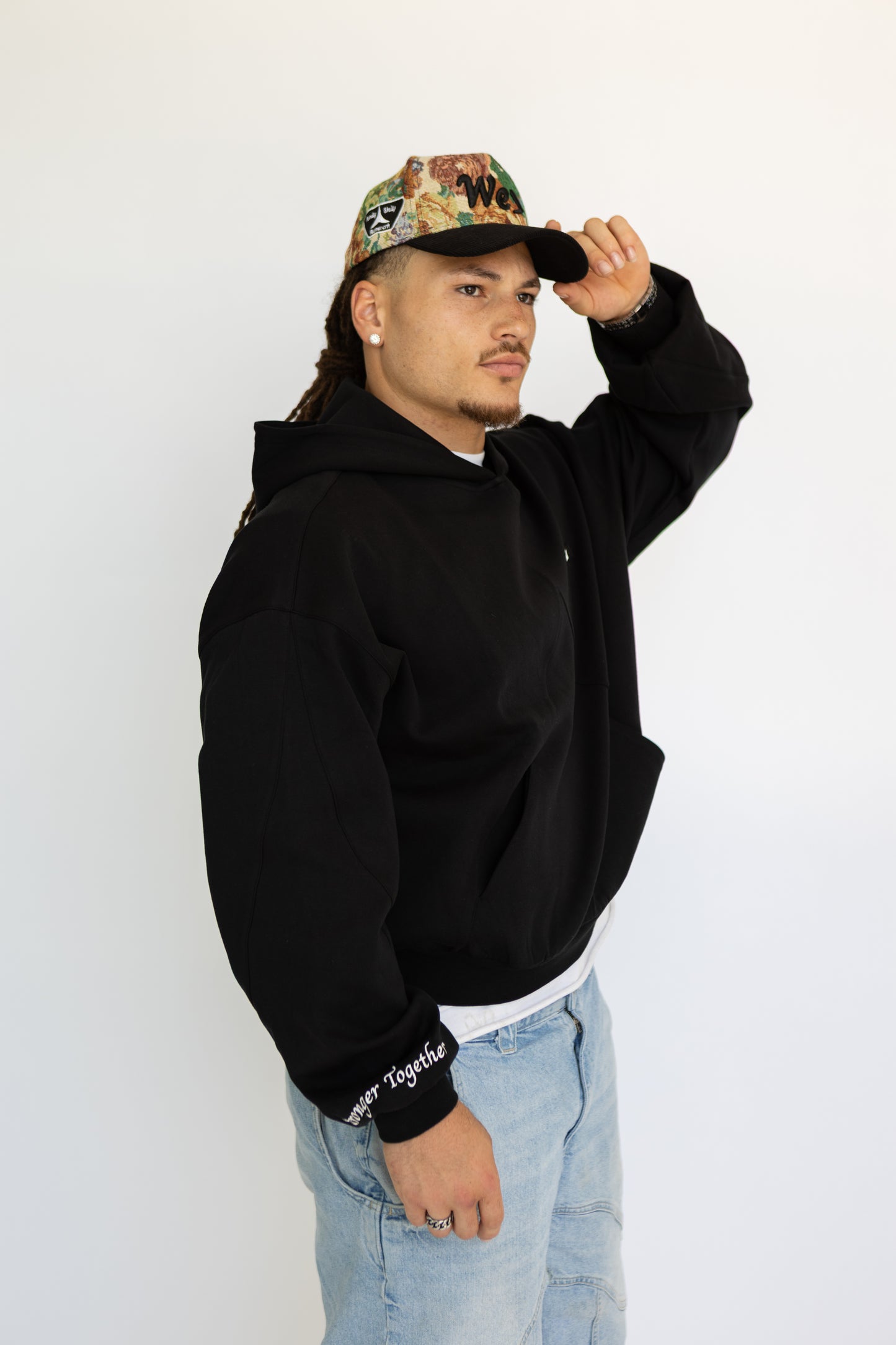 Man wearing a black hoodie, light blue jeans, and a cap with a white background
