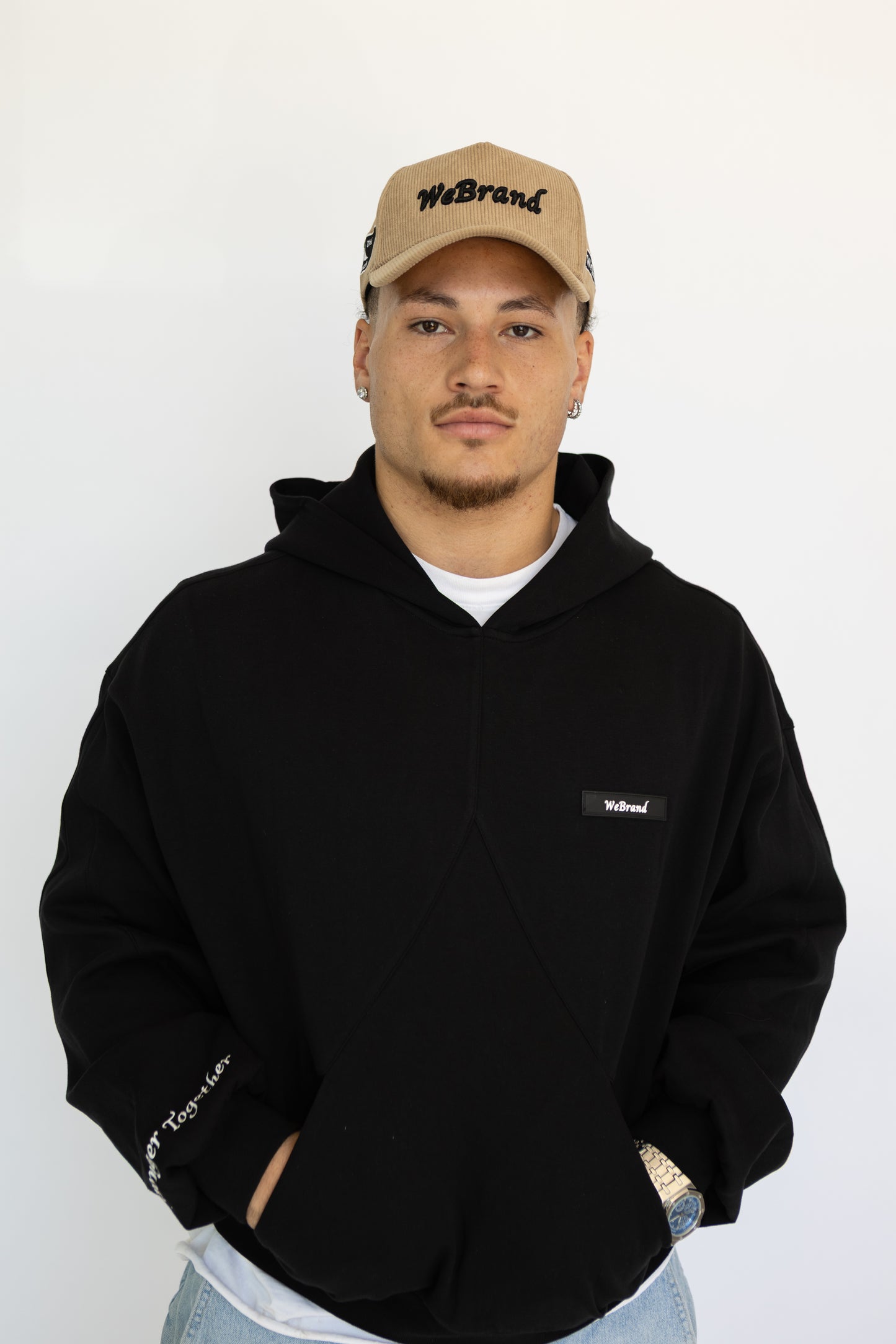 Man wearing a black hoodie and beige cap with 'WeBrand' logo on a white background