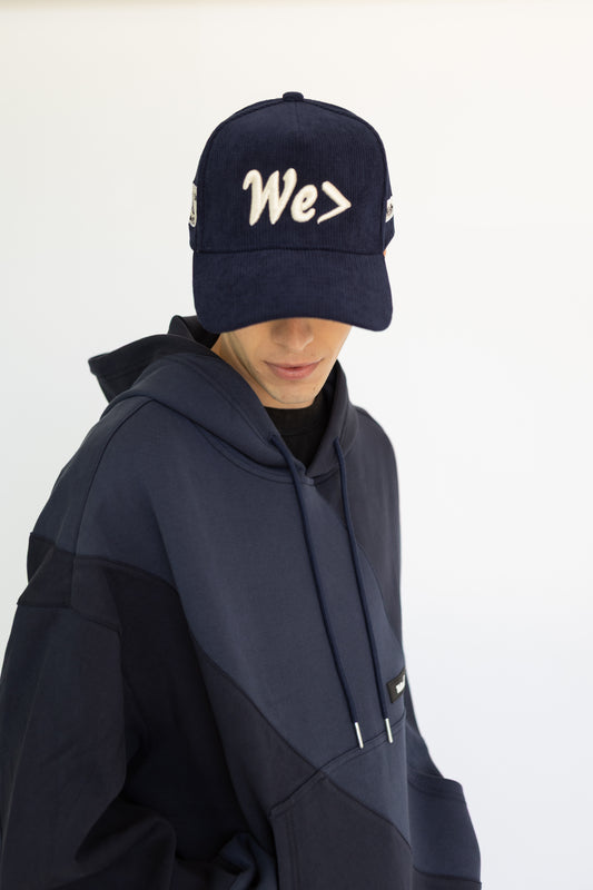 Person wearing a navy cap with 'We>' text and matching navy hoodie on a white background