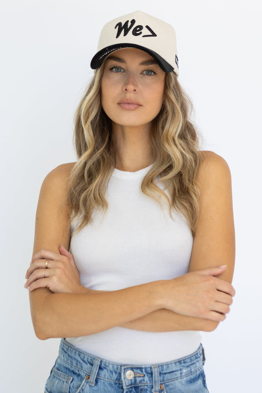 Woman wearing a white tank top, blue jeans, and a cap with 'We' logo on a white background