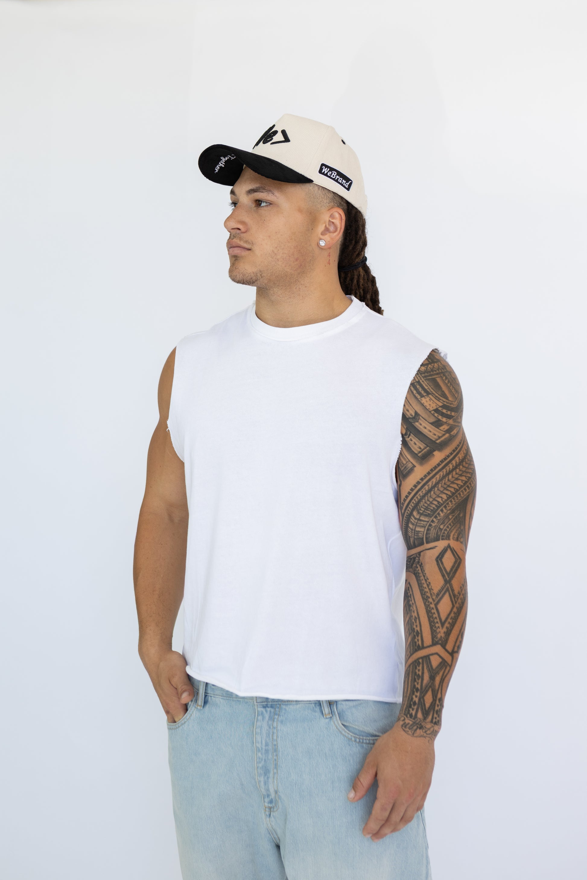 Man wearing a white sleeveless shirt, light blue jeans, and a cap on a white background