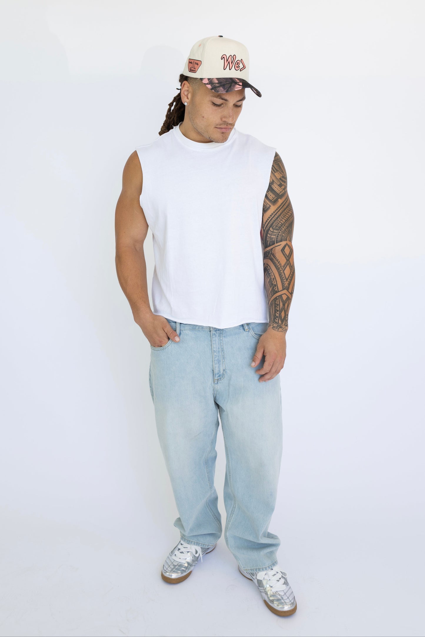 Man wearing a white sleeveless shirt, light blue jeans, and a cap on a white background