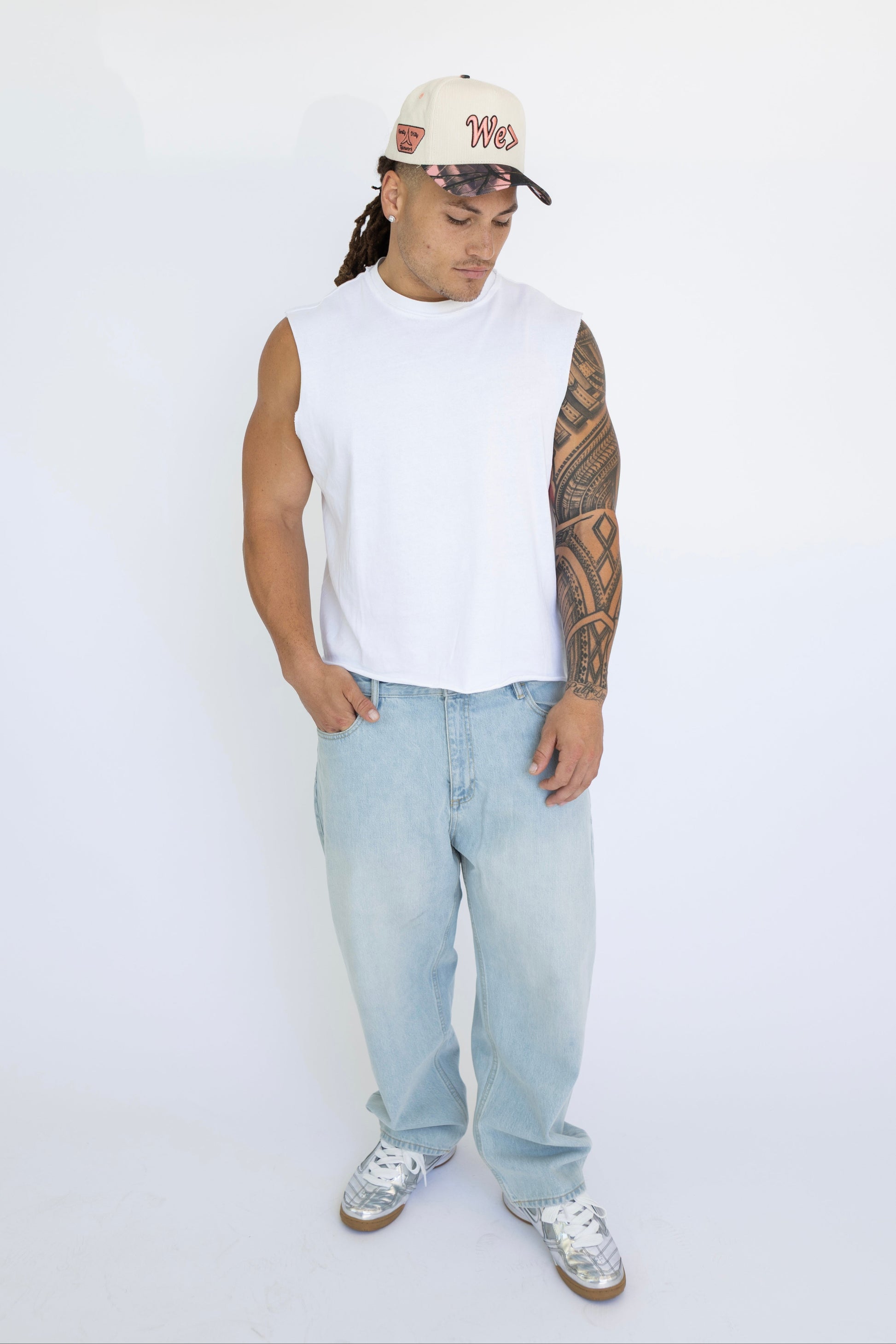 Man wearing a white sleeveless shirt, light blue jeans, and a cap on a white background