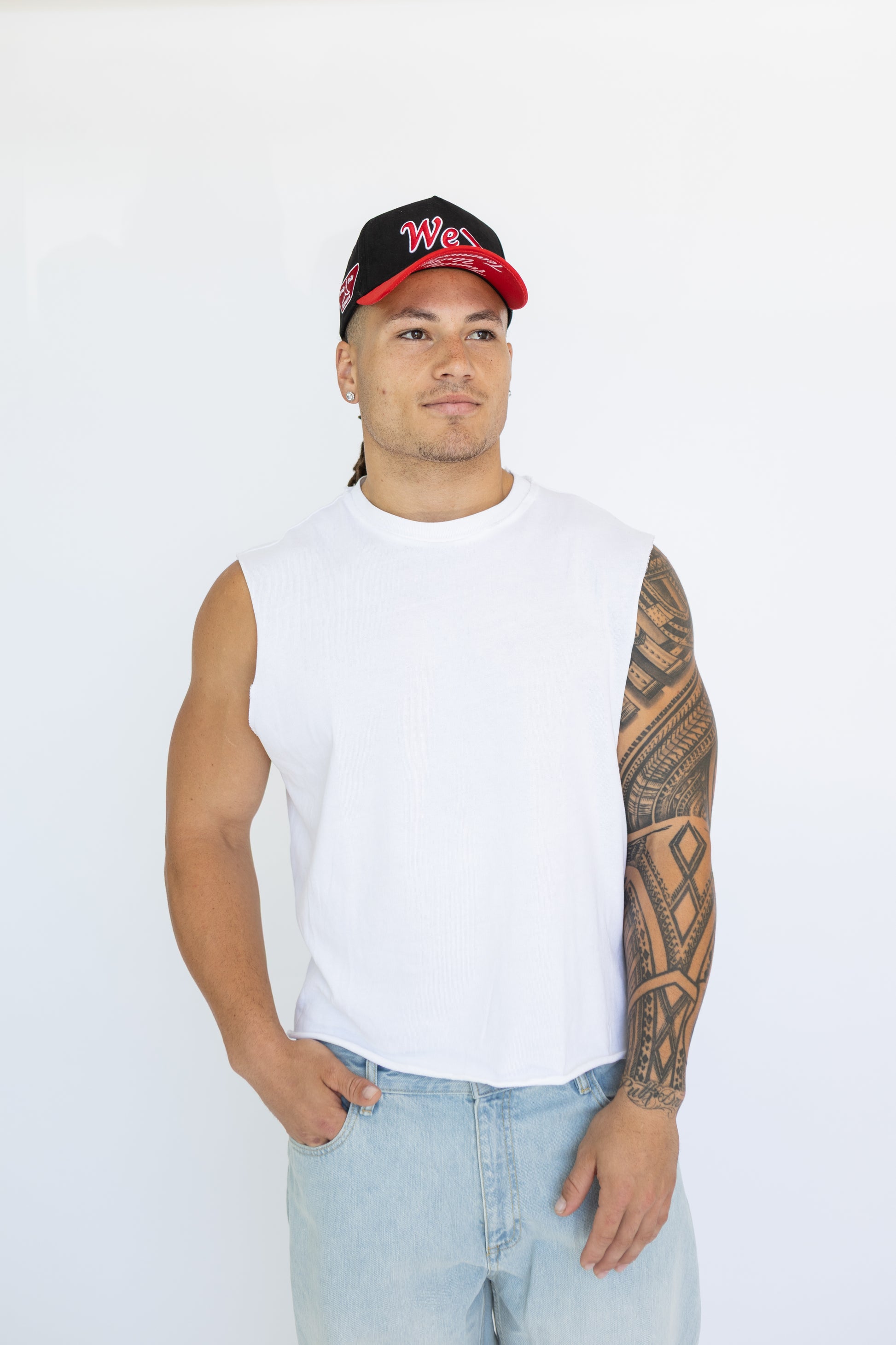 Man wearing a white sleeveless shirt, light blue jeans, and a black cap with red brim on a white background