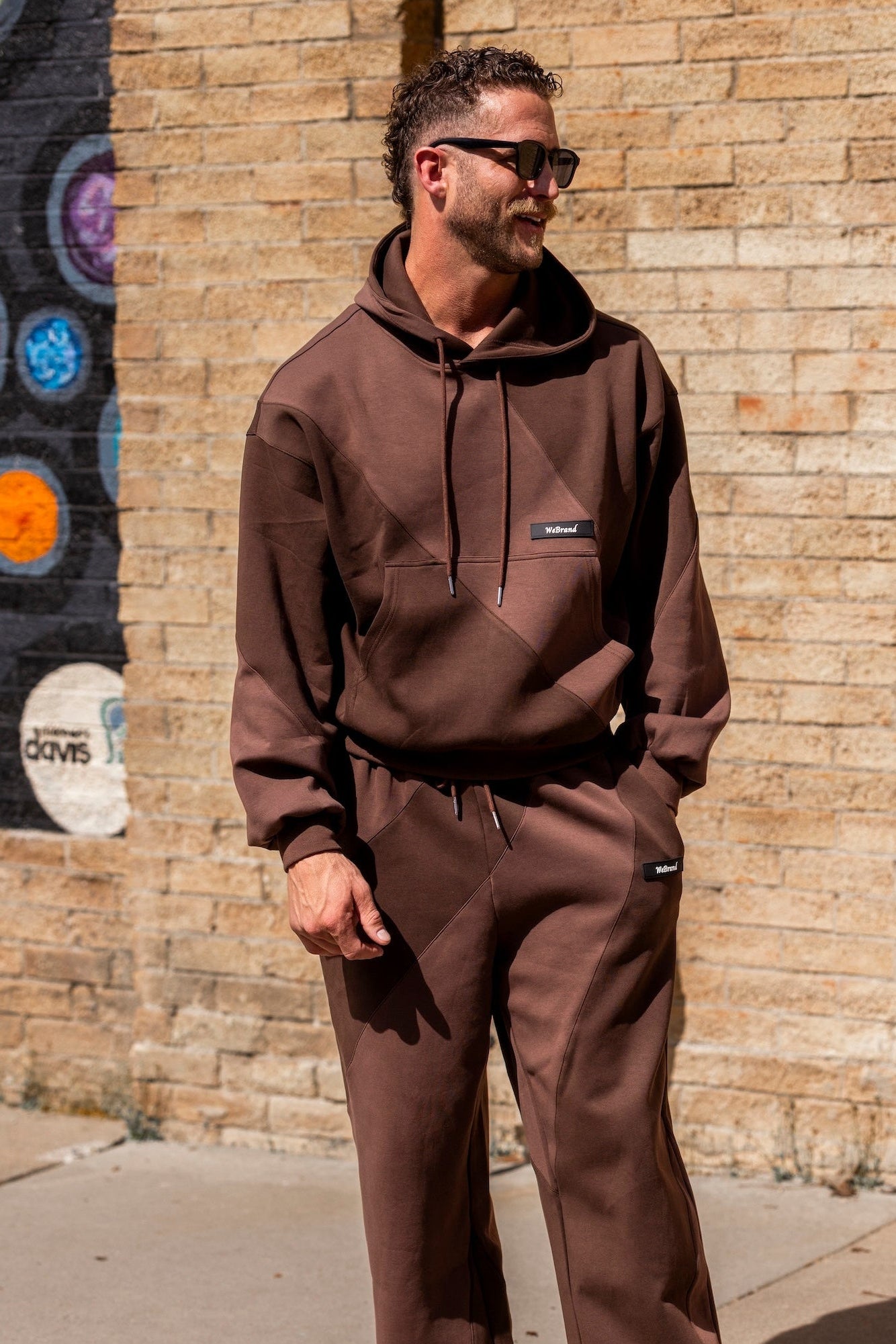 Man wearing a brown hoodie and pants standing against a brick wall with colorful abstract art.