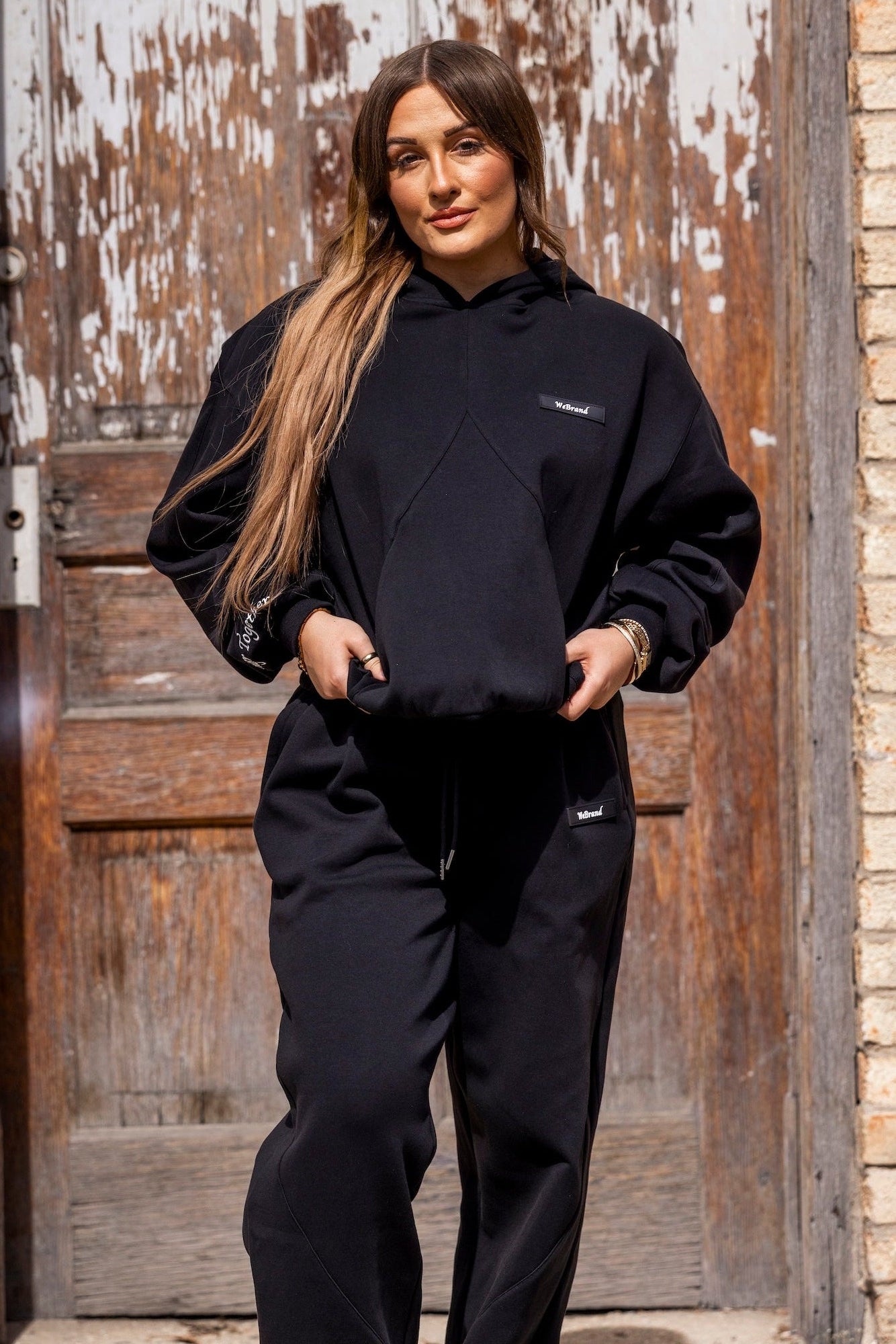 Person wearing a black hoodie and pants standing in front of a weathered wooden door.