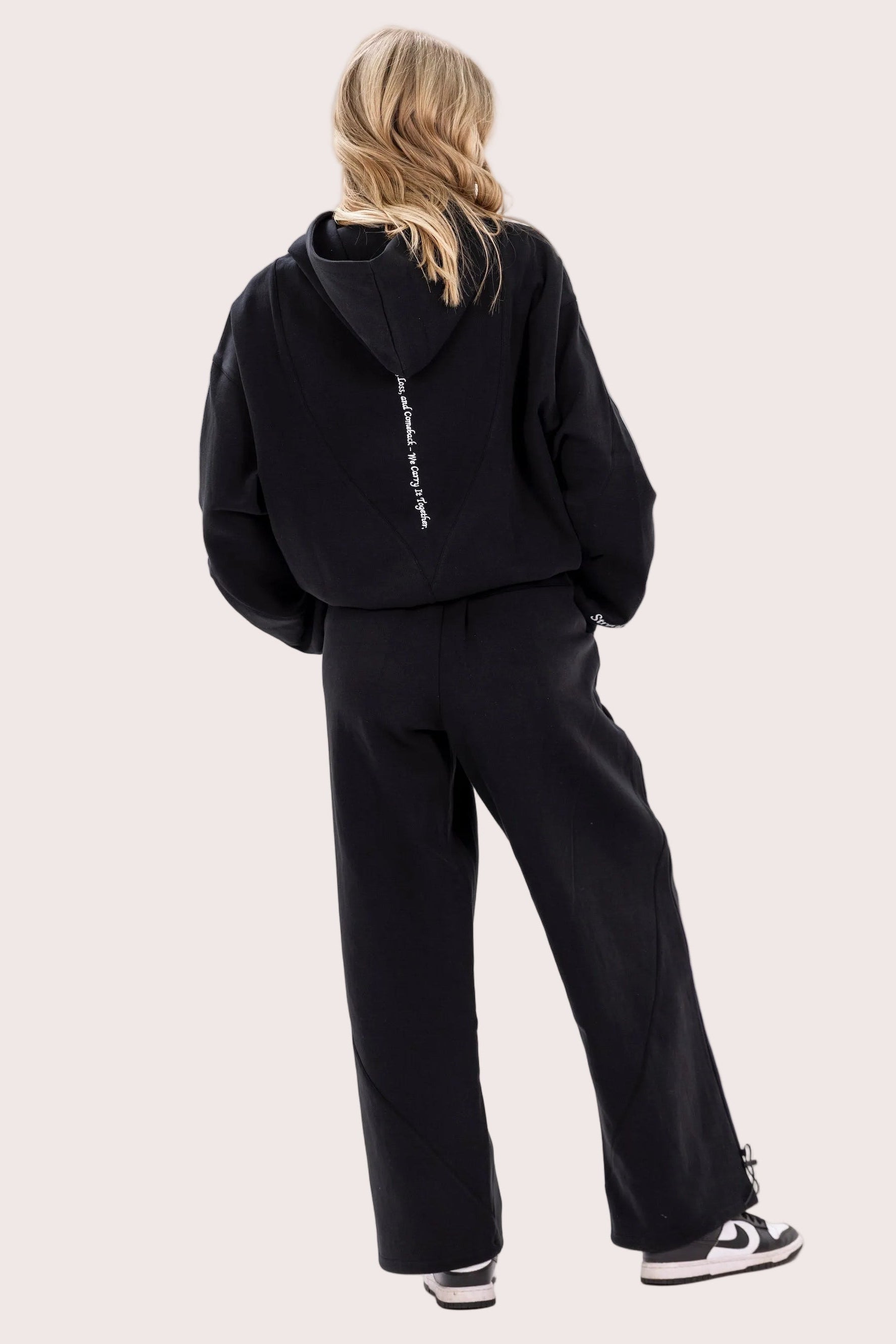 Person wearing a black hoodie and pants with a visible brand logo on a light pink background