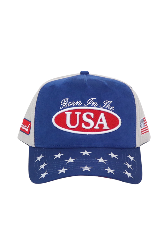 A patriotic trucker-style baseball cap with a royal blue front and brim, white mesh side panels, and a red oval patch reading 'USA' with the phrase 'Born In The' above it. The brim is decorated with white stars, and small American flag patches are visible on the side.