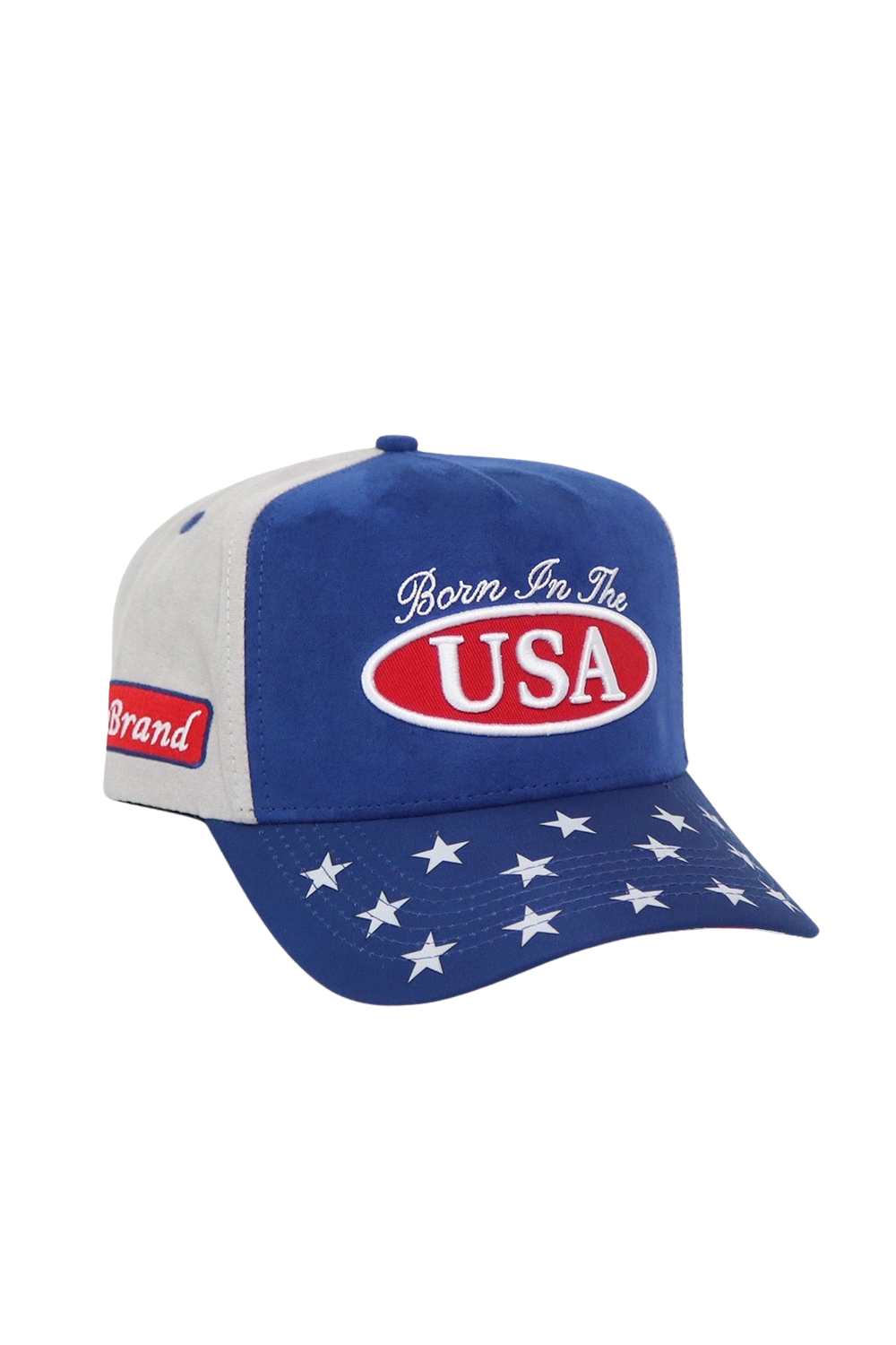 A patriotic baseball cap with a blue front and brim, white side panels, and white stars on the brim. The front features a red oval logo with 'USA' in white, and white cursive text above it reading 'Born In The'.